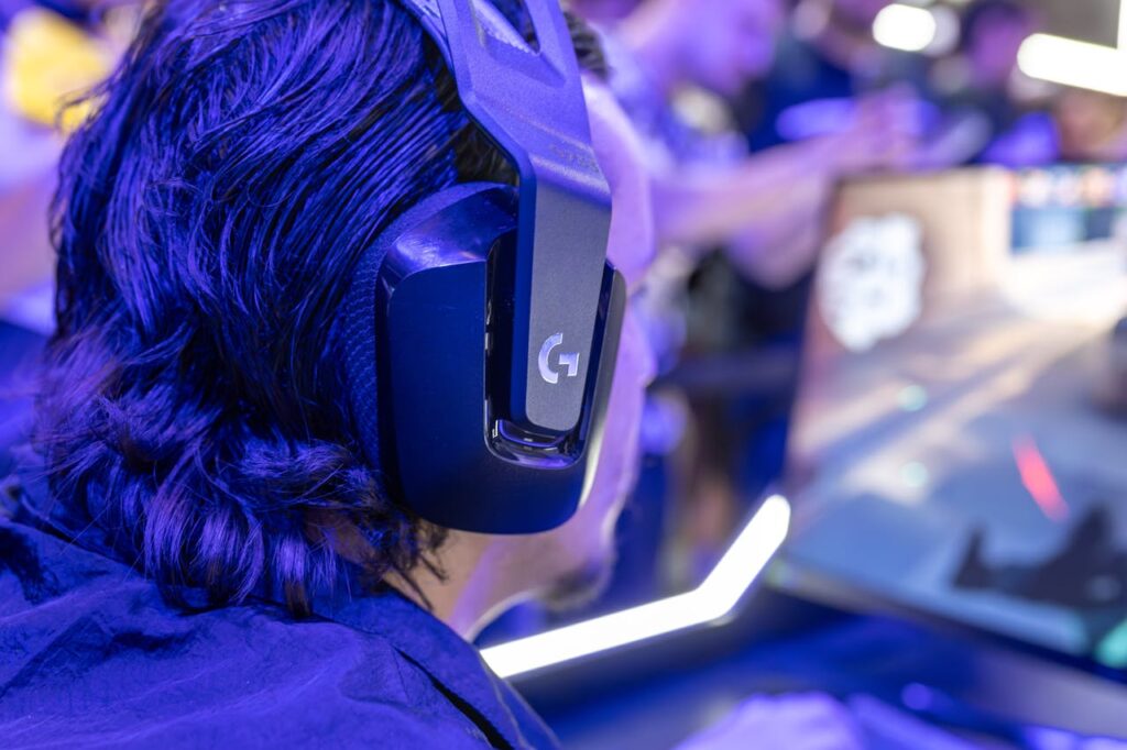 pexels photo 28993117 Focused gamer with headphones playing a video game on a PC under blue lighting in a crowded room.