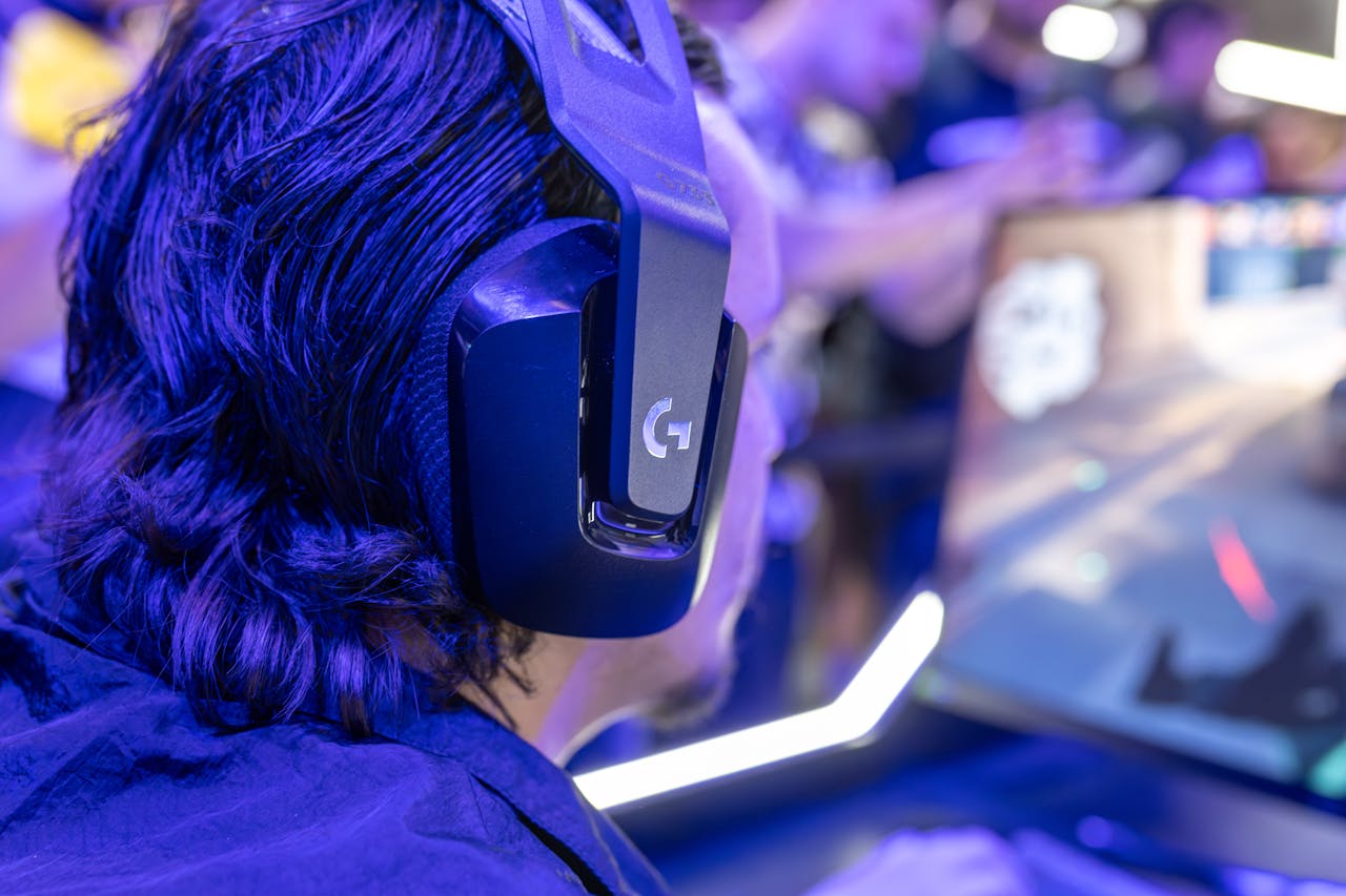 Focused gamer with headphones playing a video game on a PC under blue lighting in a crowded room.